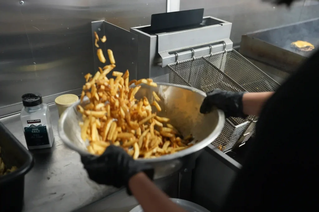 Gloved hand tossing fresh fries from a metal bowl next to the fryer